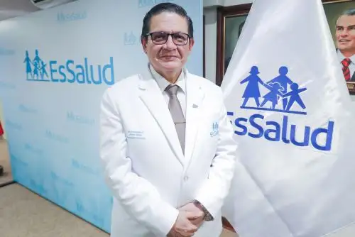 Profesional con bata junto a la bandera de EsSalud; fotografía institucional referencial.